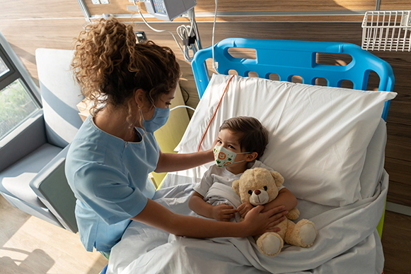 Boy lying down on hospital bed and sweet nurse comforting him both wearing facemasks